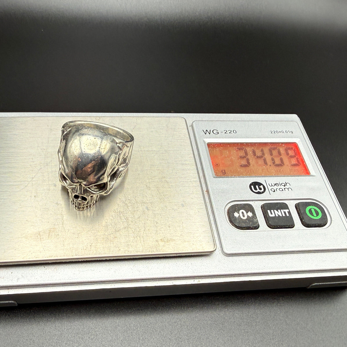 A silver skull ring displayed on a digital scale showing a weight of 34.00 grams.